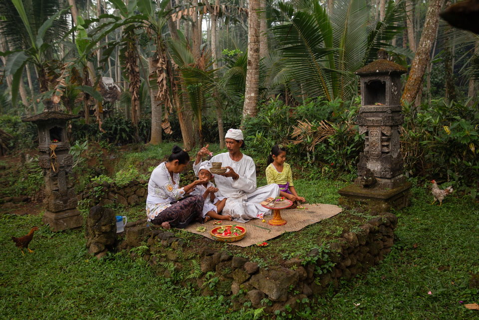 A family sits on a mat making offerings in a garden shrine.