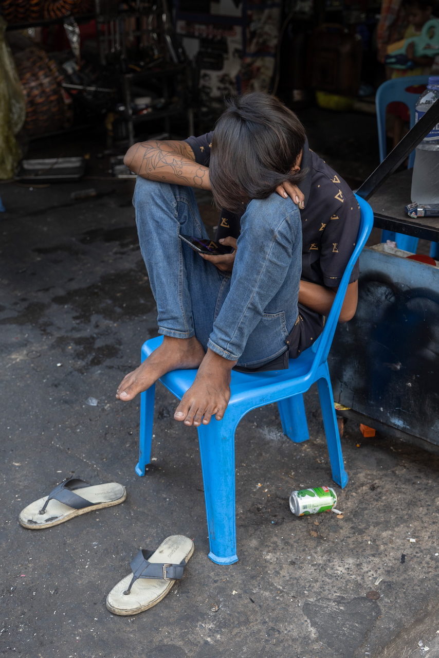 Two people share a phone on a blue plastic chair in the street.