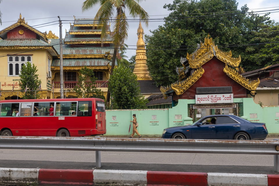 A golden pagoda and tiered temple roofs rise behind a green roadside wall as a red bus and a blue car pass.