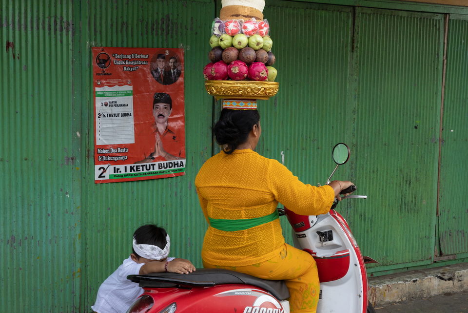 A woman in orange rides a red scooter carrying a tiered fruit offering on her head, past a green corrugated wall pasted with election posters.