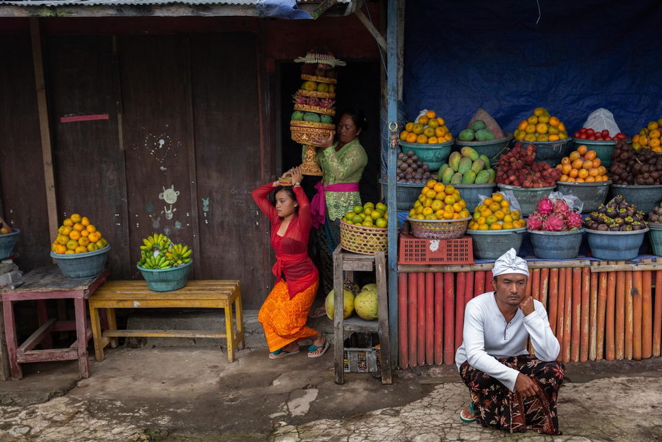 Women carry tiered offerings past a roadside fruit stall stacked with oranges and mangoes.
