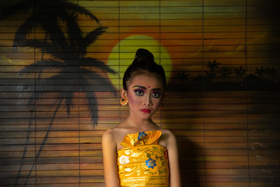 A young woman in gold against a blind painted with a palm and setting sun.