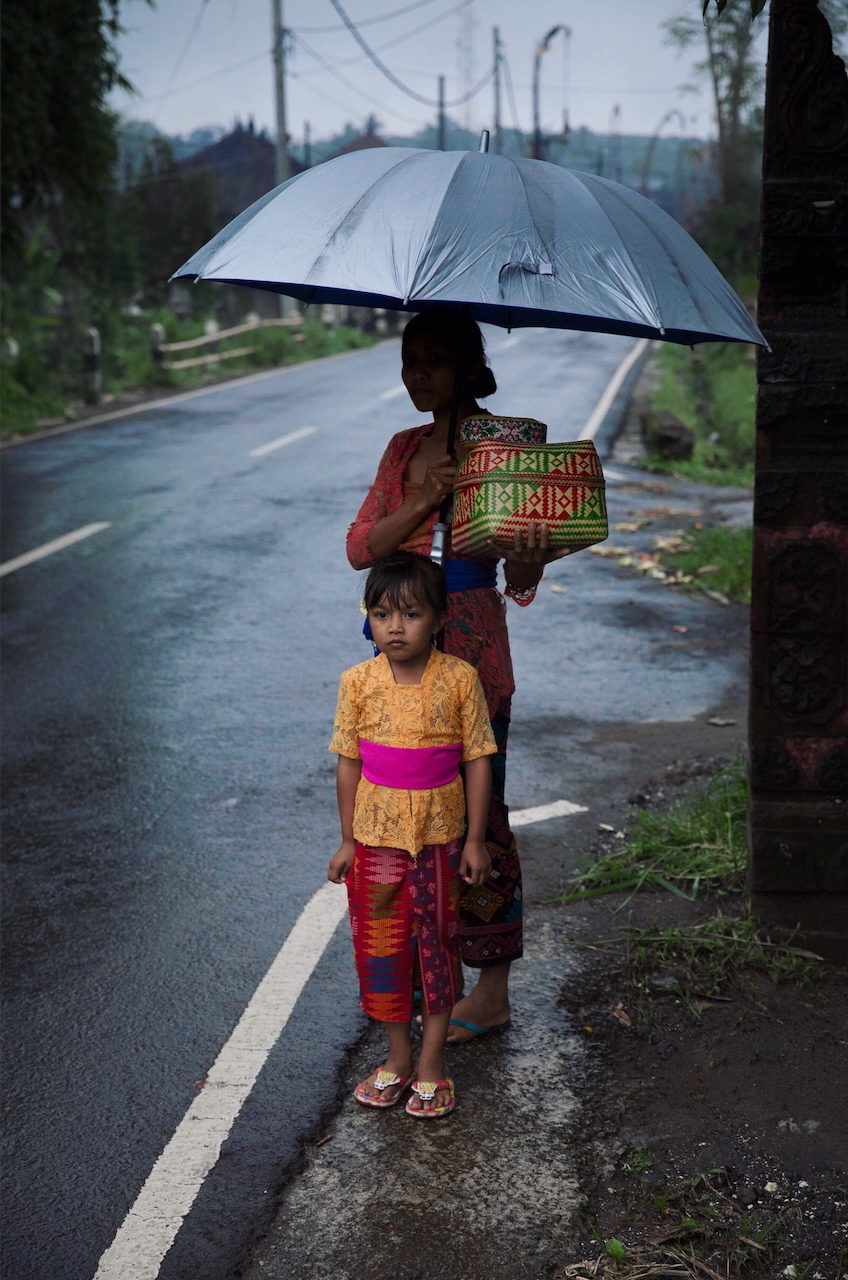 A woman and child stand on a wet road beneath an umbrella.