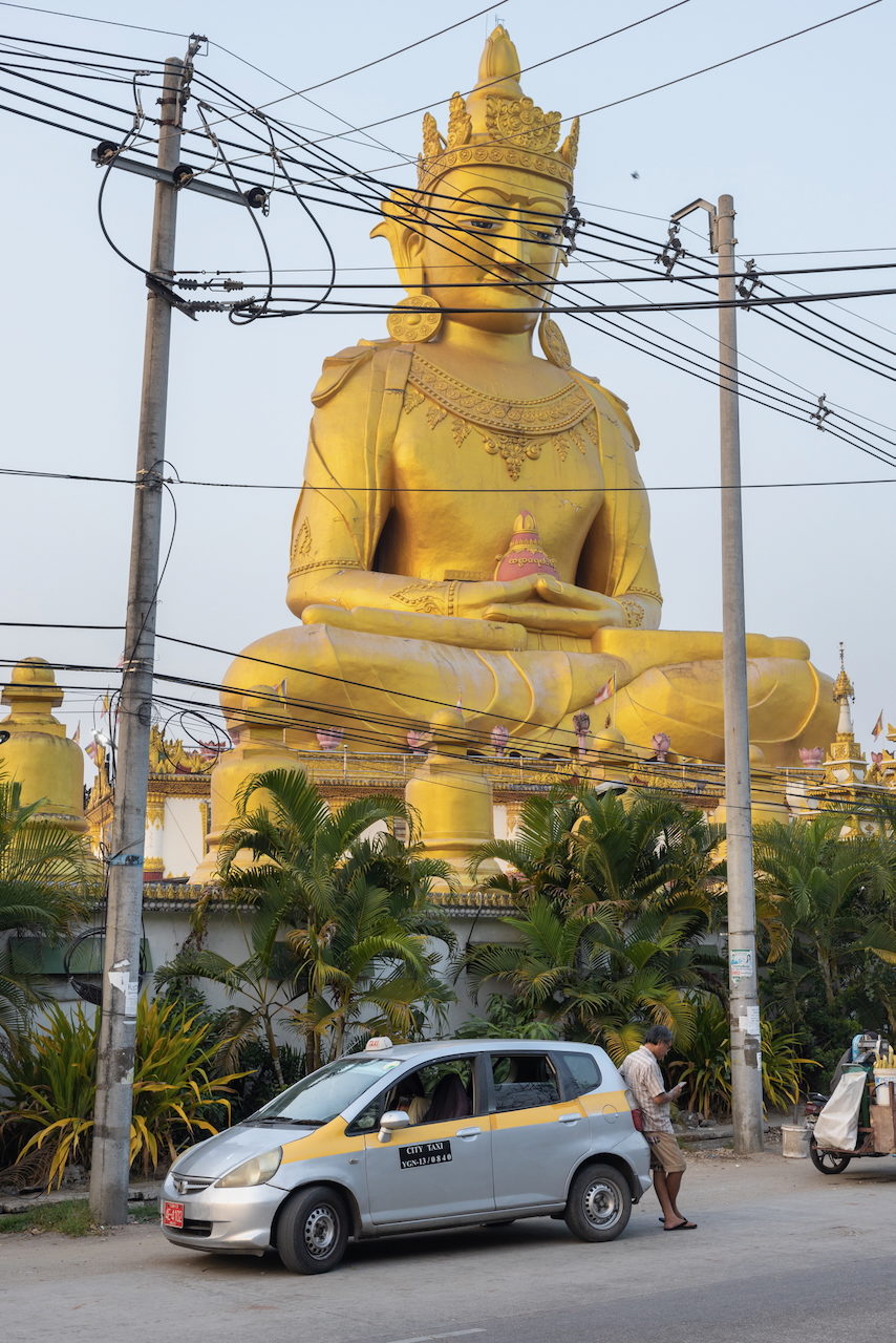 A large golden Buddha rises behind palm trees and power lines.
