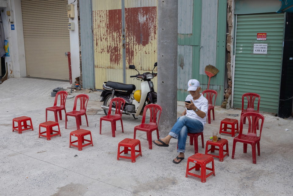 Red plastic stools set out on a street, a man seated among them.