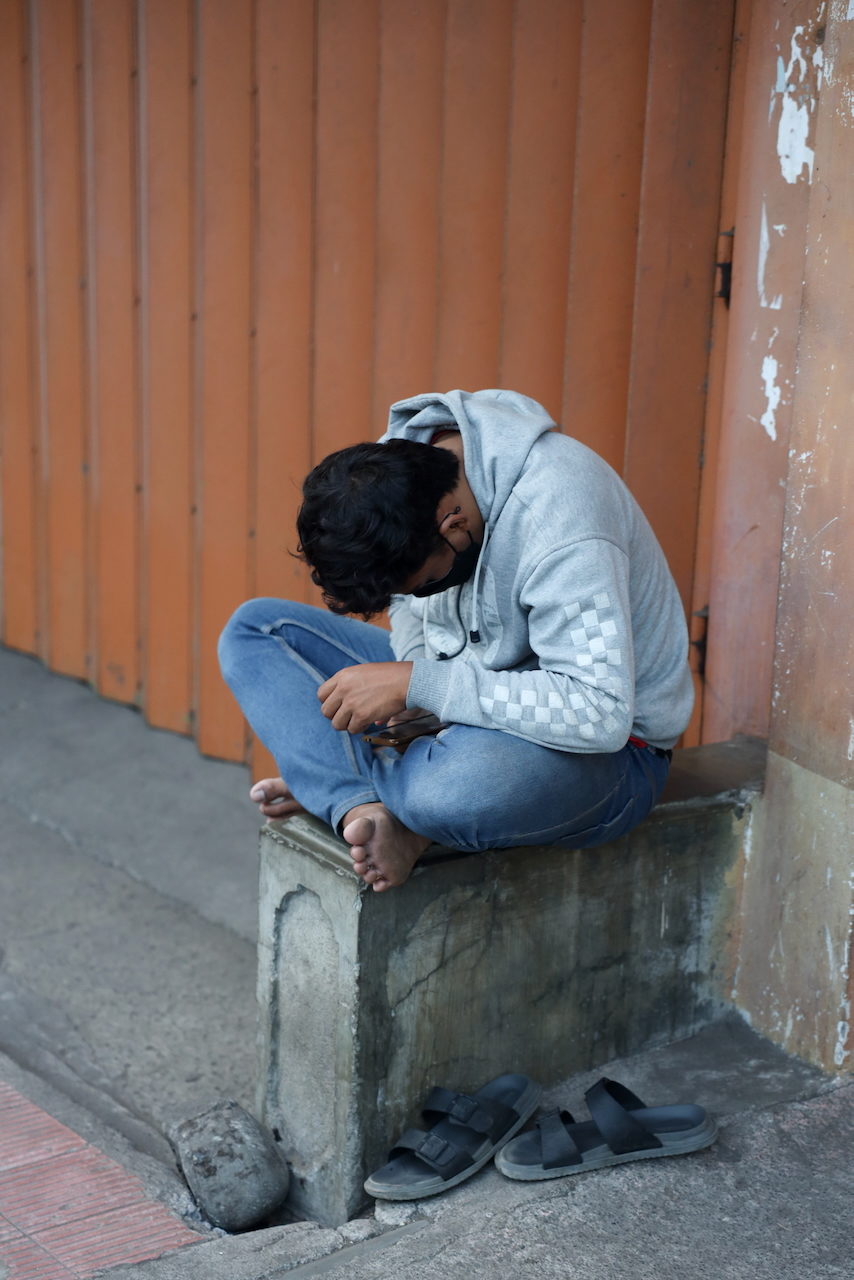 A young man in a hooded top sits on a kerb absorbed in his phone.