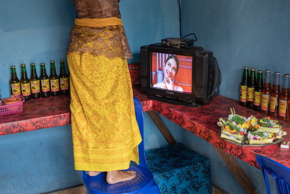 A woman in a gold sarong stands beside a television on a red cloth.