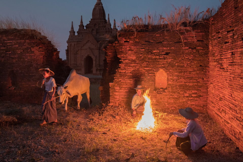 Villagers tend a fire beside a brick temple wall at dusk, a white ox standing in the glow.