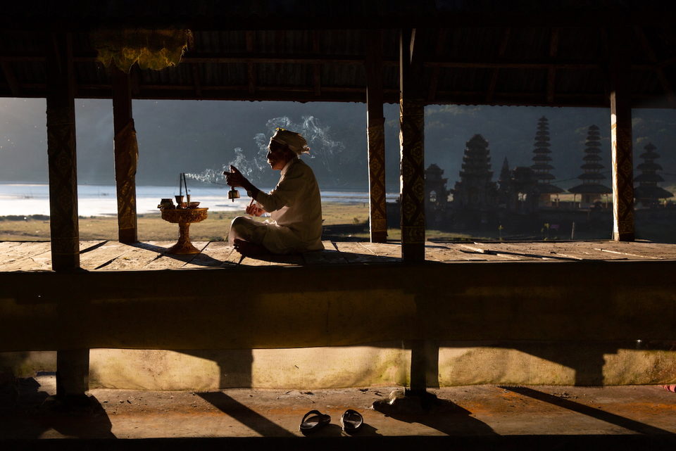 A priest sits in silhouette with incense, sea and temple beyond.