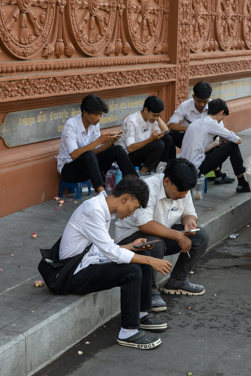 Students in white shirts sit on steps, most of them looking at phones.