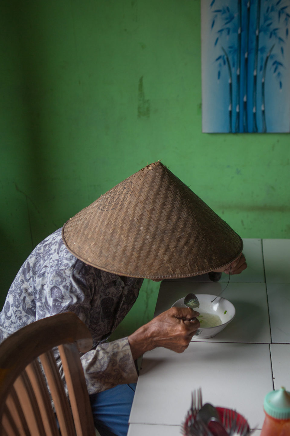 A woman in a conical hat spoons food into a bowl against a green wall.