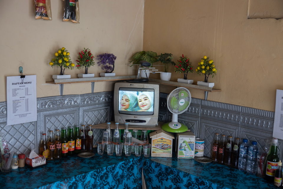A television on a shelf of plastic flowers above rows of bottled drinks.