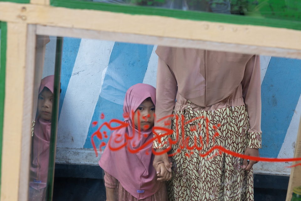 Two women in headscarves stand against a wall painted with pale blue stripes and red calligraphy.