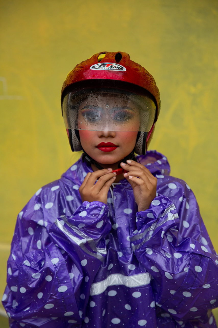 A dancer in full makeup fastens a red helmet over a purple polka-dot rain poncho.