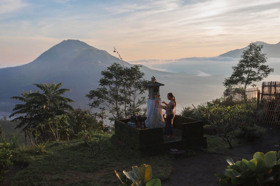 A woman makes an offering at a hilltop shrine at dawn, volcanoes and mist beyond.