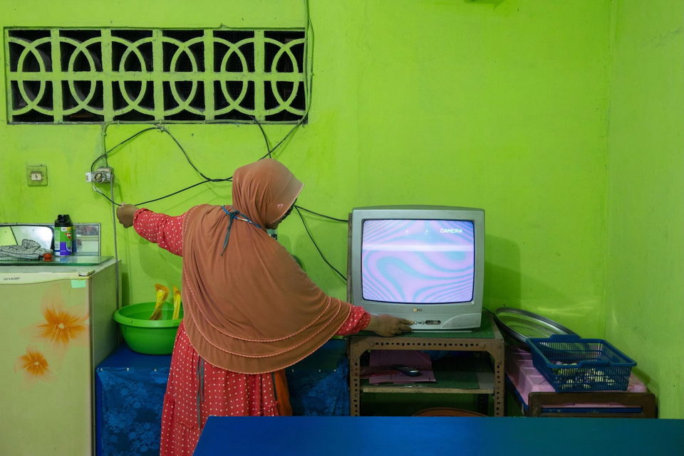 A woman in a headscarf adjusts a television against a green wall.