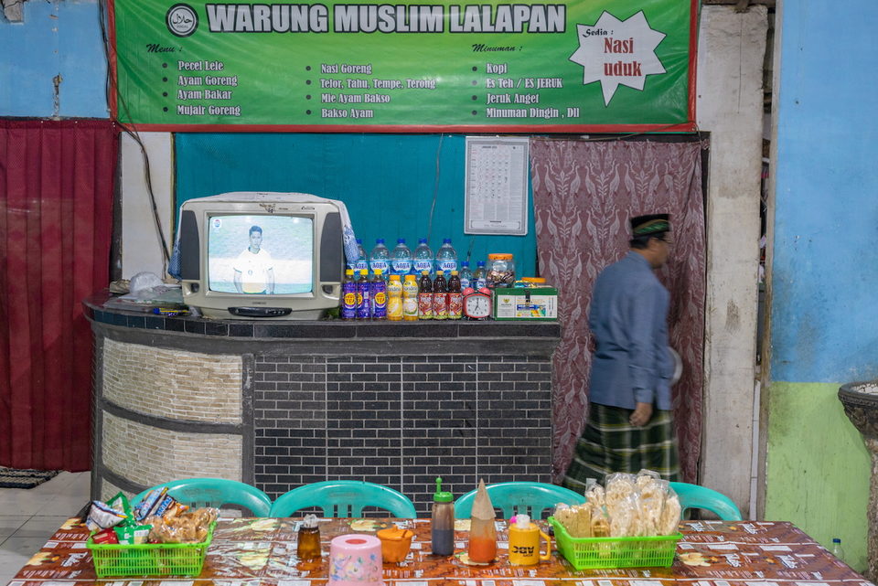 A warung counter under a green menu banner, a television among the bottles.