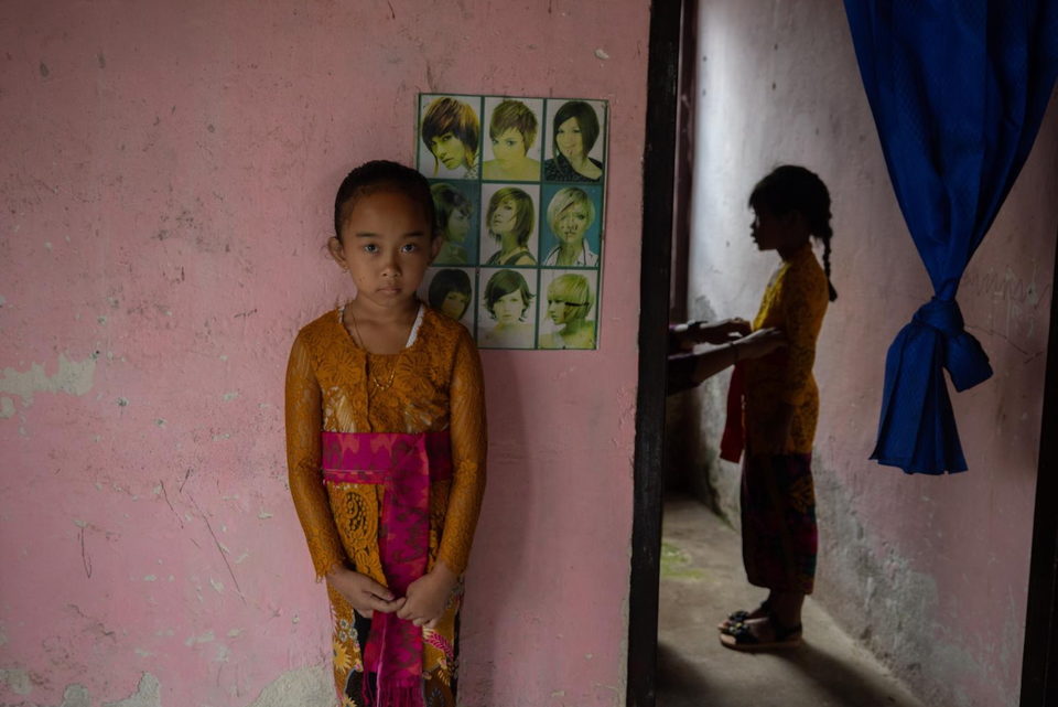 A girl in gold and pink stands against a pink wall hung with hairstyle posters.