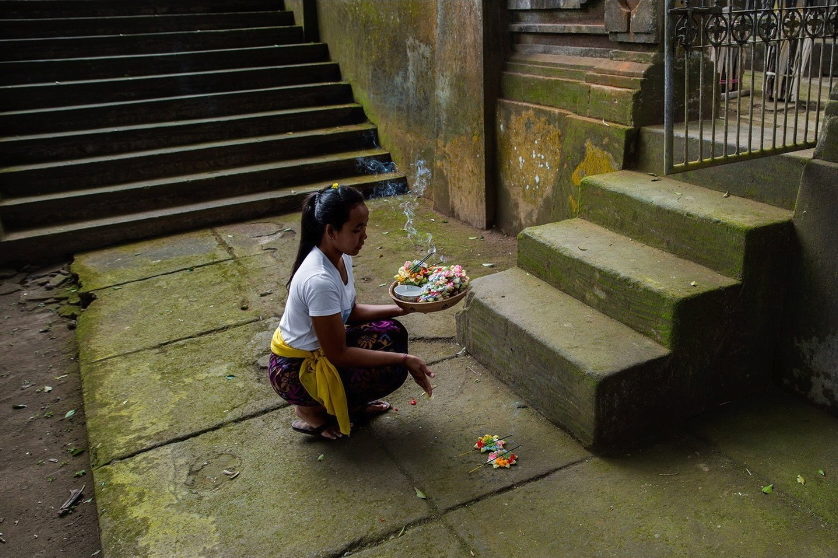 A woman crouches with an offering tray at the foot of stone steps.
