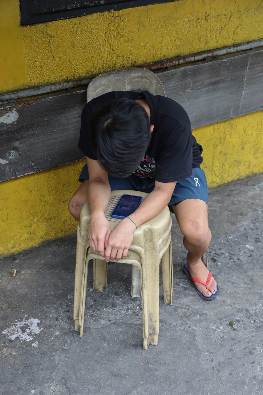 A young man sits hunched on a plastic stool, head down over a tablet.