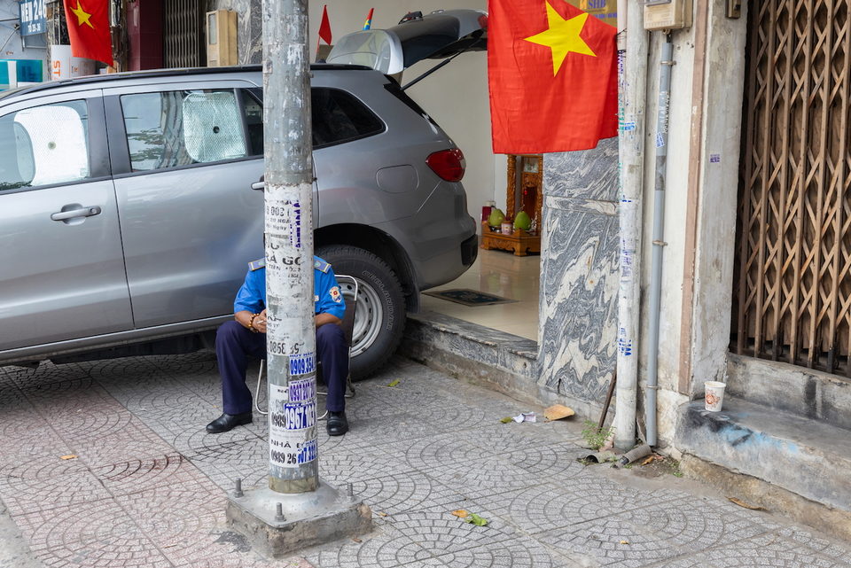A man sits against a pole beside a parked car and a Vietnamese flag.