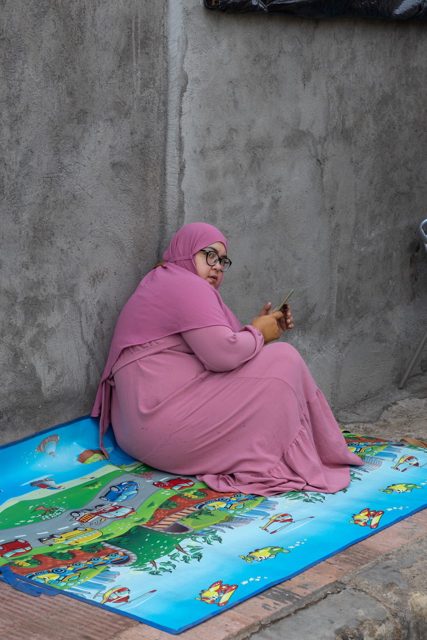 A woman in pink sits cross-legged on a printed mat holding her phone.