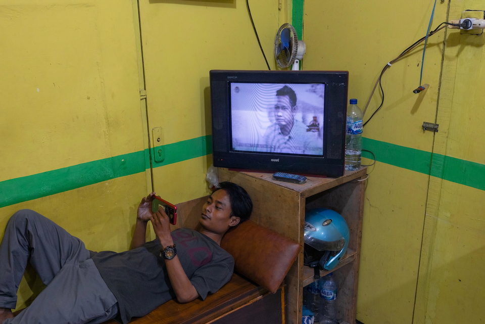 A man reclines on a bench watching television against a yellow and green wall.