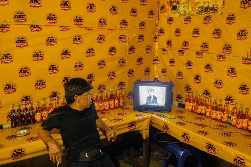 A man in a black cap watches television in a warung papered with yellow packets.