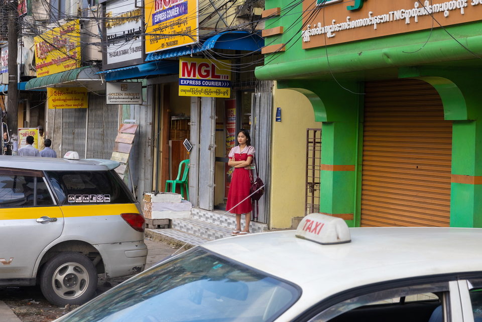 A woman in a red dress stands in a doorway on a street of yellow and green shopfronts, above a white taxi.